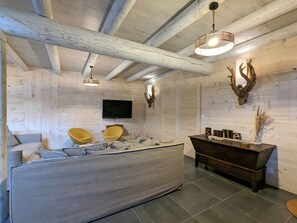 Een rustiek interieur met een groot bed, twee gele stoelen, een houten bankje, een aan de muur bevestigde televisie en een plafond met zichtbare balken.