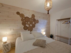 Een slaapkamer met een houten hoofdbord, een bed met wit beddengoed, een decoratieve wanddecoratie, een hanglamp en een kledingkast met spiegel en kleding.