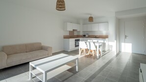 A modern living space with a sofa, a dining table with chairs, a kitchen area, and a hanging light fixture. - Modern apartment with parking - 4 guests (Mimizan)