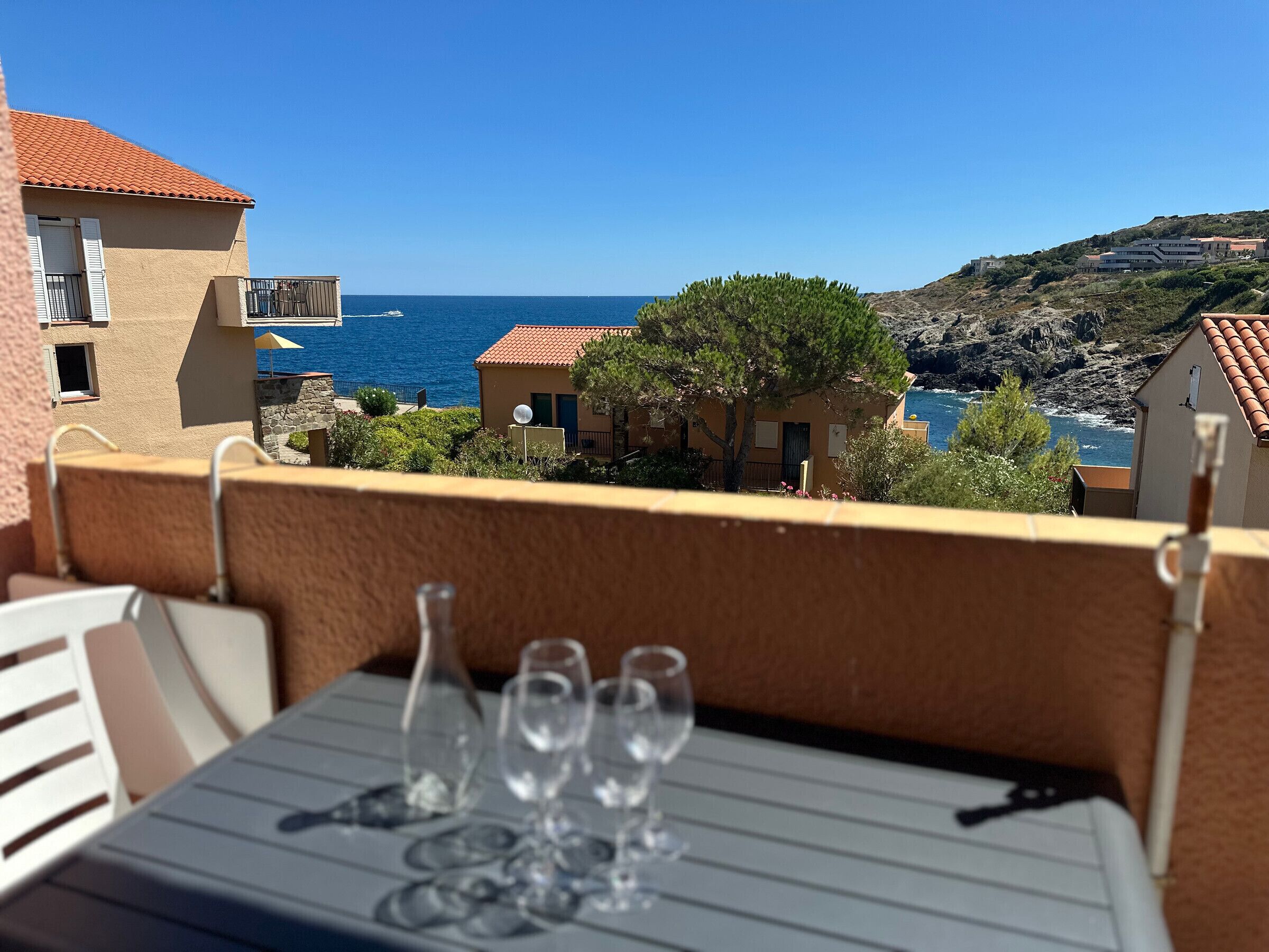 A balcony with a table set for dining, overlooking a coastal landscape with buildings and the sea.