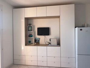A compact kitchen with a built-in TV, a white refrigerator, and a shelf with decorative items. - Charming sea view T2 with private parking (Collioure)