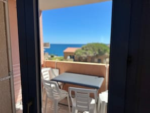 An open door leading to a balcony with a table and chairs, overlooking a coastal area with buildings and a clear sky. - Charming sea view T2 with private parking (Collioure)