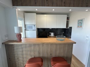 A modern kitchen with a wooden countertop, white cabinets, a microwave, a sink, and a coffee machine. There are two red chairs at the counter. - Apartment, sea view, pets welcome - 4 guests (Cabourg)