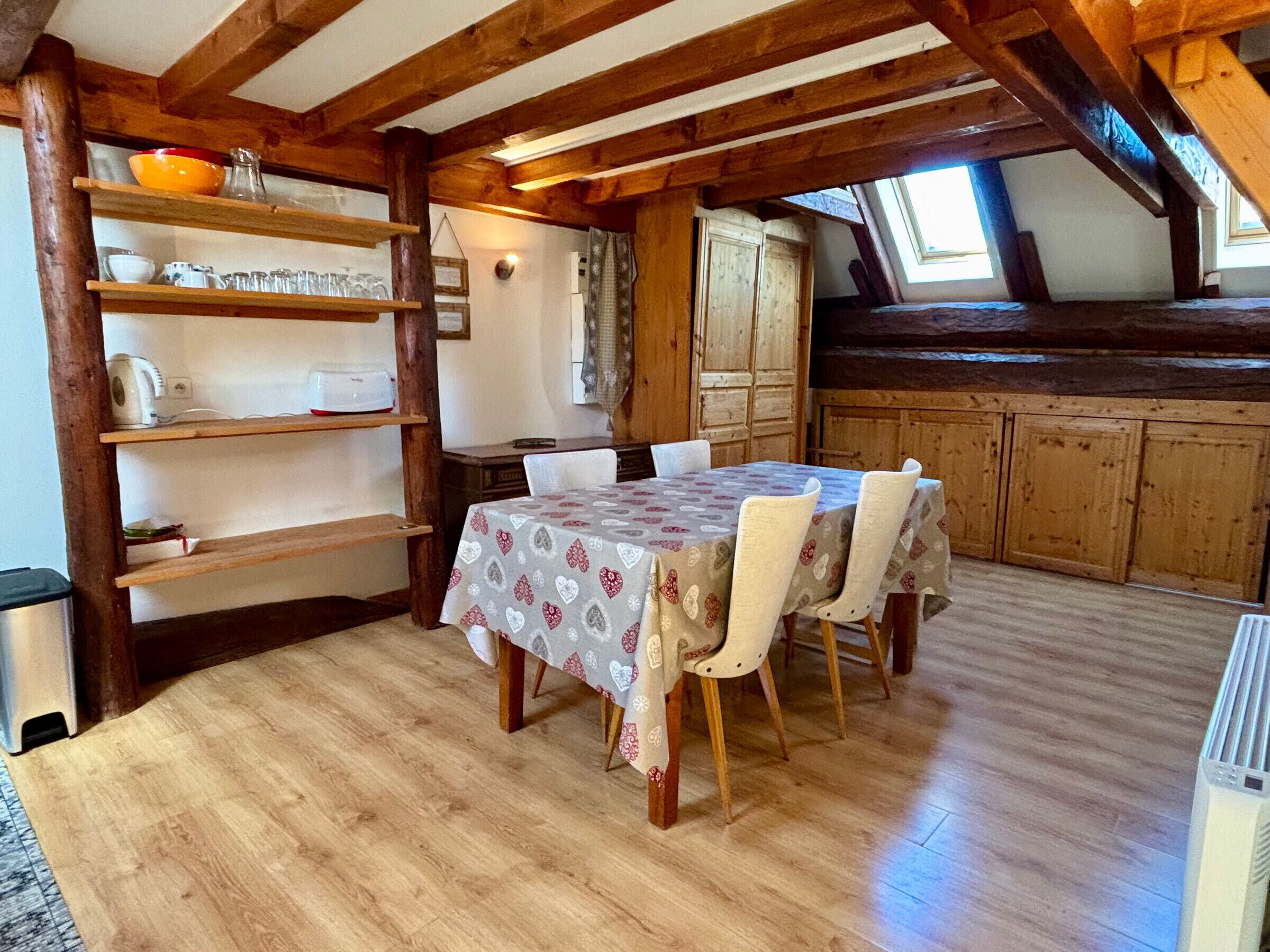 Un coin repas avec une table dressée pour quatre personnes, des poutres en bois au plafond et un espace cuisine avec des placards en bois.