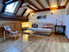 Ein gemütliches Zimmer mit Holzbalkendecke, einem Sofa mit gemusterten Kissen, einem kleinen Tisch und einem Stuhl.