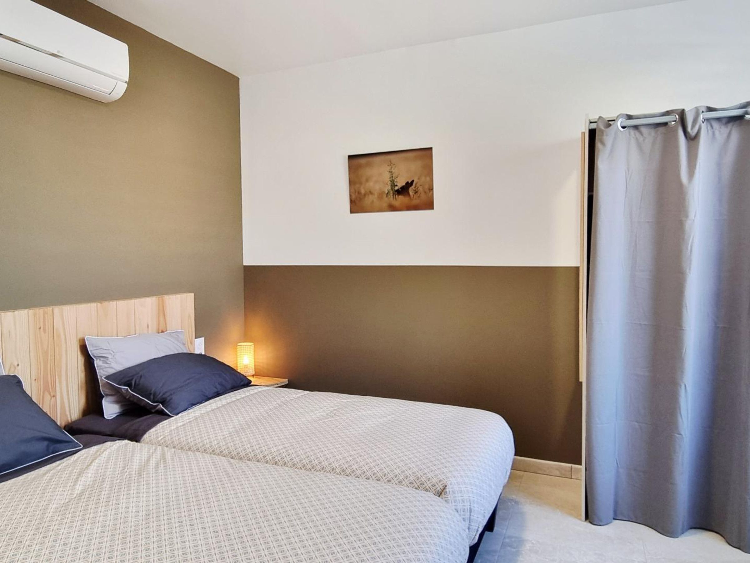 Ein Hotelzimmer mit zwei Betten, einem Kopfteil, einem Nachttisch mit Lampe, einer Wandklimaanlage und einem Vorhang.