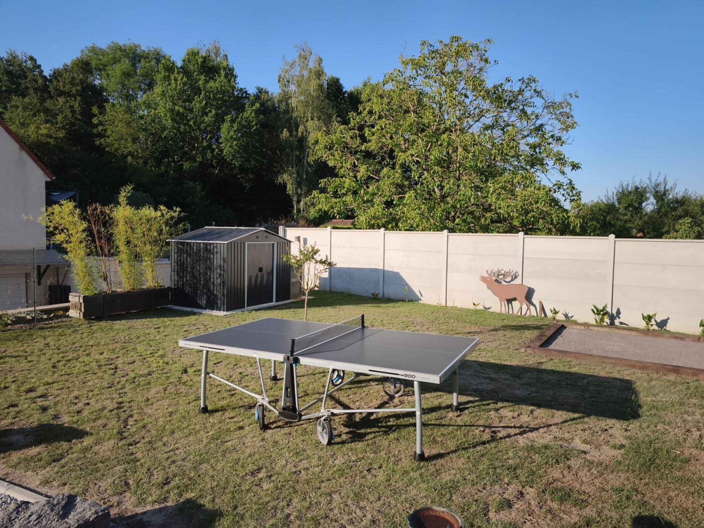 Een tafeltennistafel in een achtertuin met een tuinhuis en een hertenbeeld aan de muur.