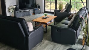 Ein modernes Wohnzimmer mit einer Sofagarnitur, einem Couchtisch, einem Fernseher und einem großen Fenster mit Blick auf Grünflächen.