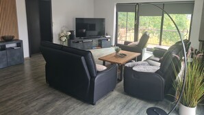 Ein modernes Wohnzimmer mit einem Flachbildfernseher, schwarzen Sofas, einem Holztisch und einem Balkon mit Blick auf Grünflächen.
