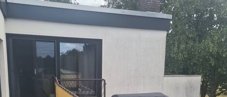 Ein überdachter Grill auf einer Holzterrasse, eine Glastür mit Schiebeöffnung zu einer überdachten Terrasse und ein Ziegelkamin.