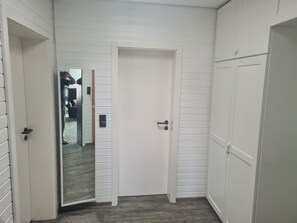 Un couloir aux murs recouverts de panneaux blancs, une porte fermée et un miroir qui reflète une pièce dont le sol est gris.
