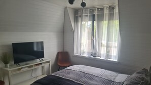 Ein Schlafzimmer mit einem Bett, einem Fernseher auf einer weißen Kommode, einem Sessel und einem Fenster mit transparenten Vorhängen.