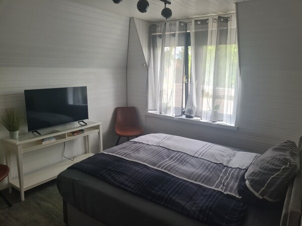 A bedroom with a bed, a TV on a white dresser, a chair, and a window with sheer curtains.