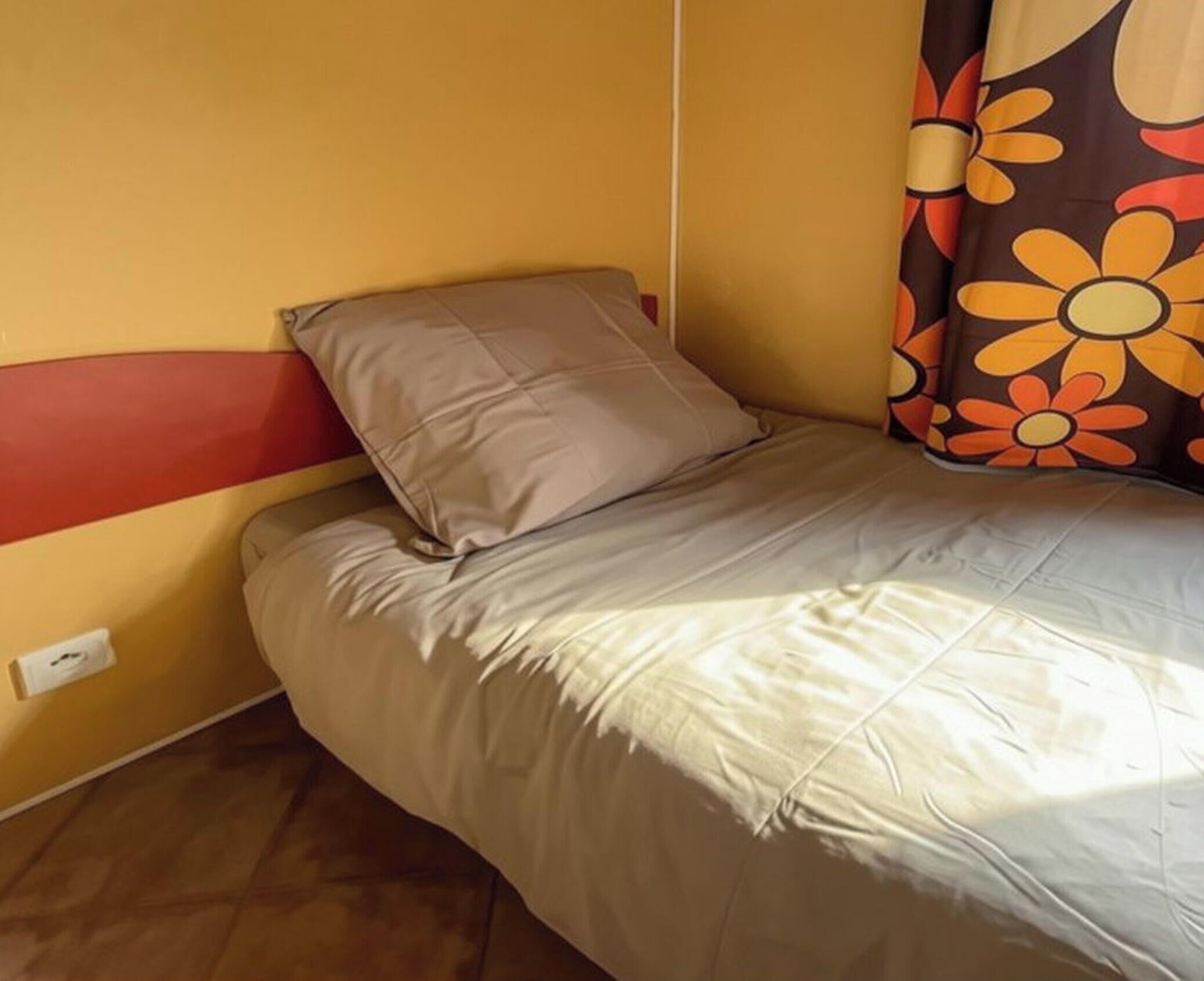 A single bed with a headboard, a pillow, and a blanket in a room with a plain wall and a power outlet.
