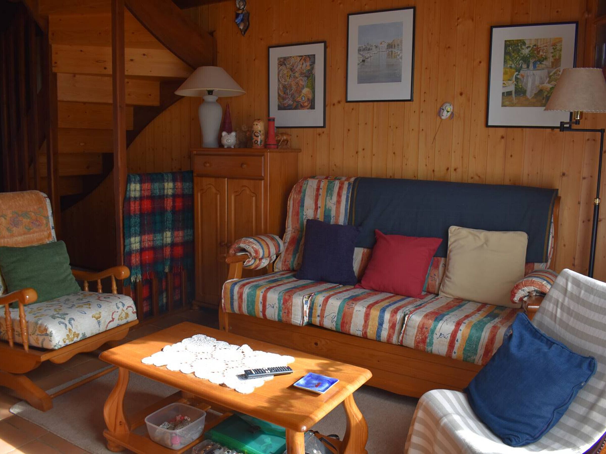 A cozy living room with a wooden staircase, a sofa with colorful cushions, a small table with a remote control, and framed artwork on the wall.