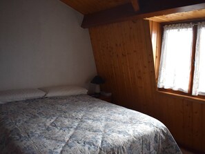 Ein Schlafzimmer mit Bett, Kissen, einem Nachttisch, einer Lampe und einem Fenster mit Spitzenvorhängen.