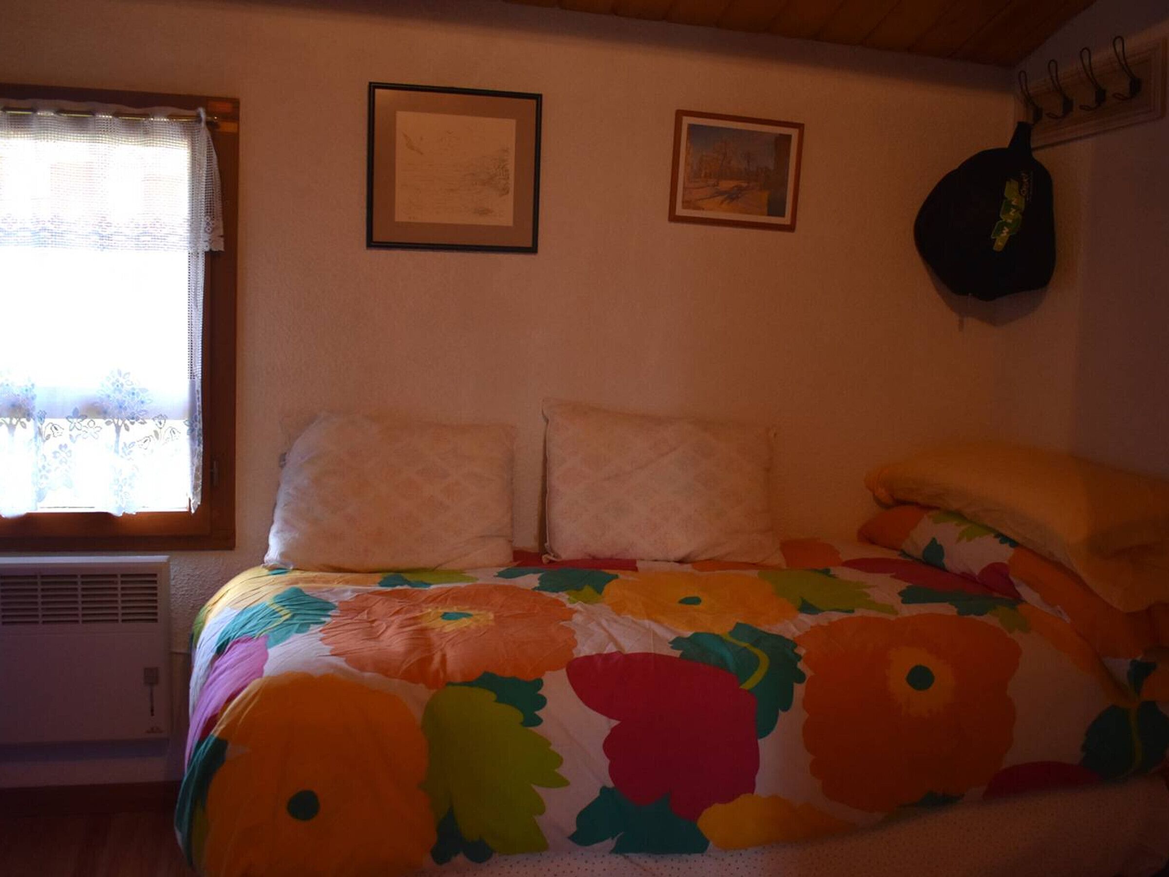Un dormitori amb un llit, dos quadres al mur, una finestra amb cortina i un objecte penjant a la paret.