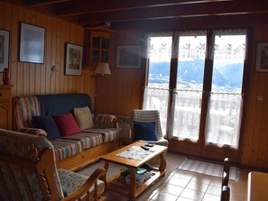 A cozy living room with a wooden interior, a sofa, a coffee table, and a view of a mountainous landscape through a large window. - Chalet with fireplace, Pyrenees view - 5 guests (Font-Romeu-Odeillo-Via)