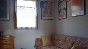 A room with a patterned sofa, a wooden dresser, a window with lace curtains, and wall decorations including posters. - Chalet with fireplace, Pyrenees view - 5 guests (Font-Romeu-Odeillo-Via)