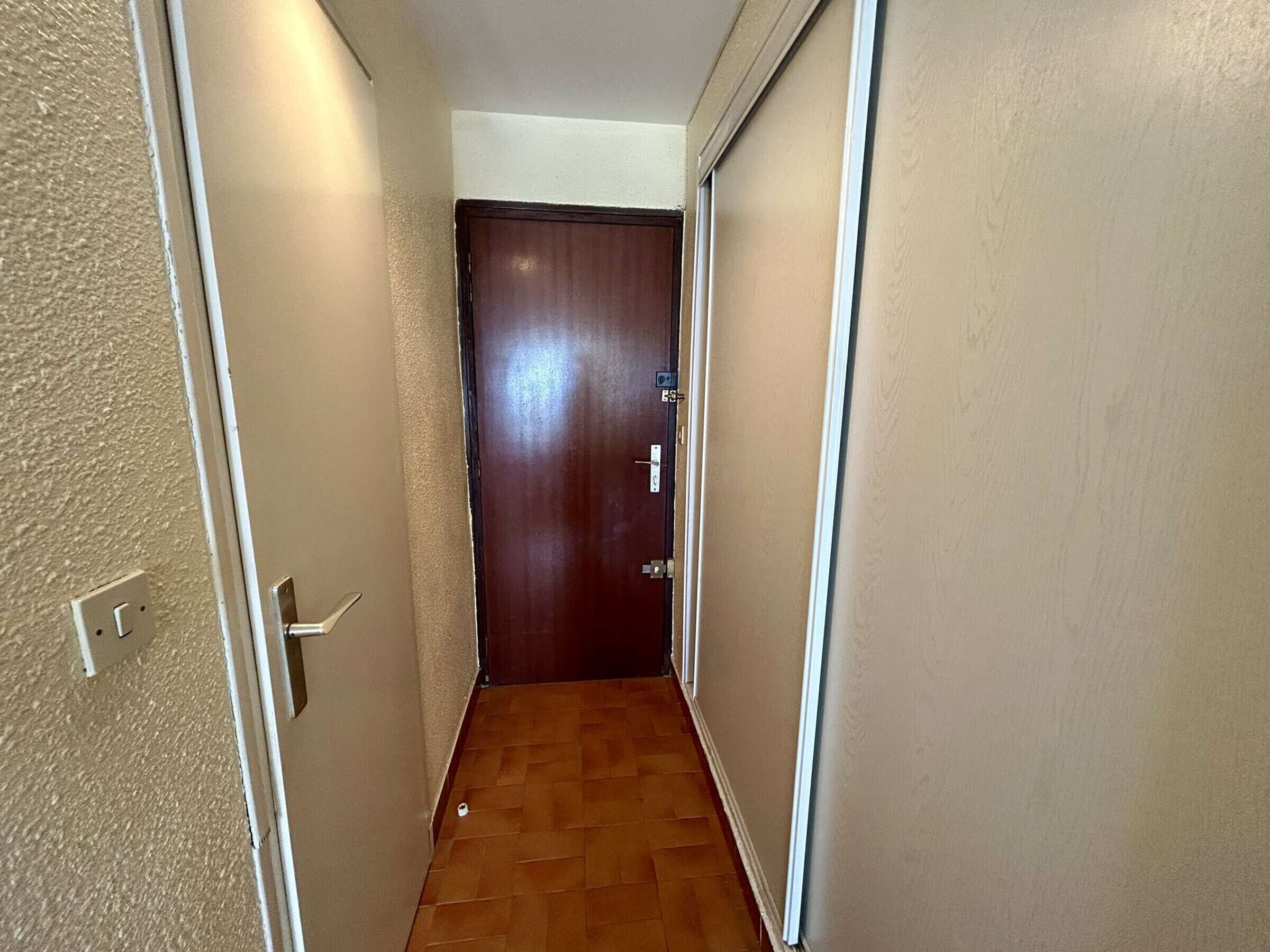 A narrow hallway with a wooden door at the end, a light switch on the left, and a white wall on the right.
