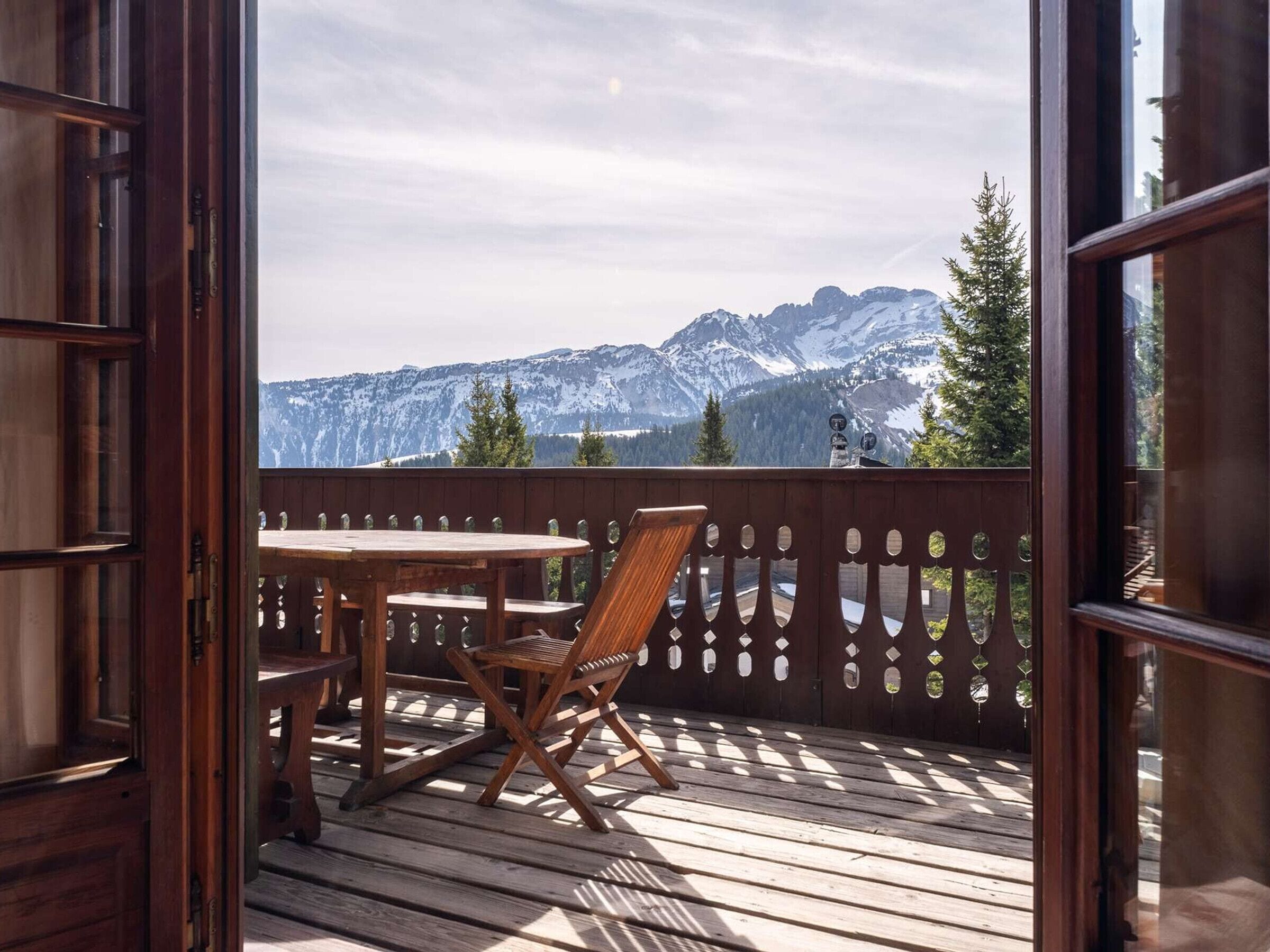 An open wooden door leading to a balcony with a table and chair, overlooking a mountainous landscape.