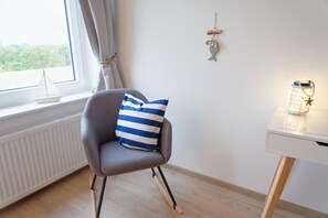 Ein Zimmer mit einem grauen Sessel, einem gestreiften Kissen, einem kleinen Tisch mit einer Laterne und einem Fenster mit Vorhängen.