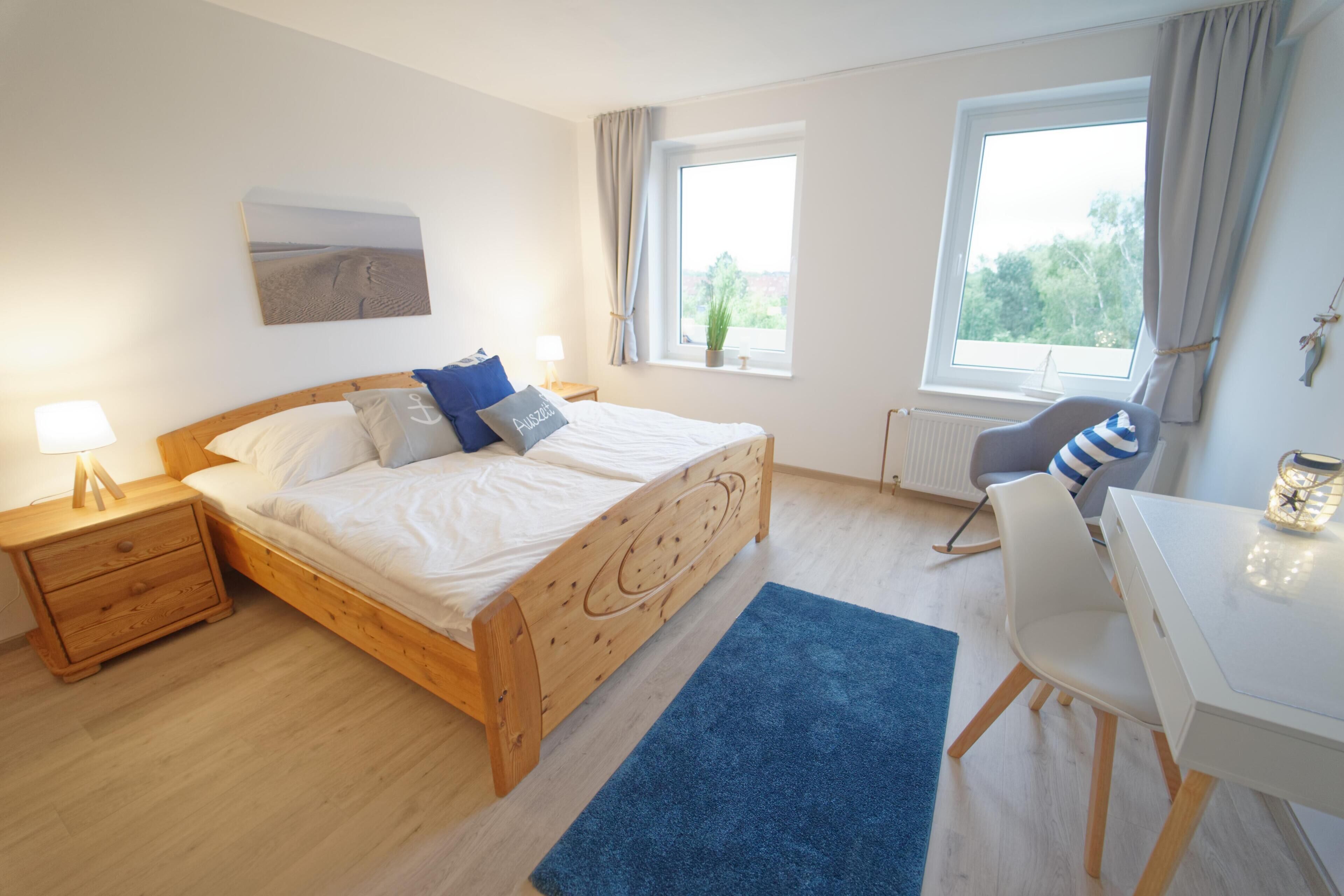 A bedroom with a wooden bed, bedside table, lamp, and a view of greenery through the window.
