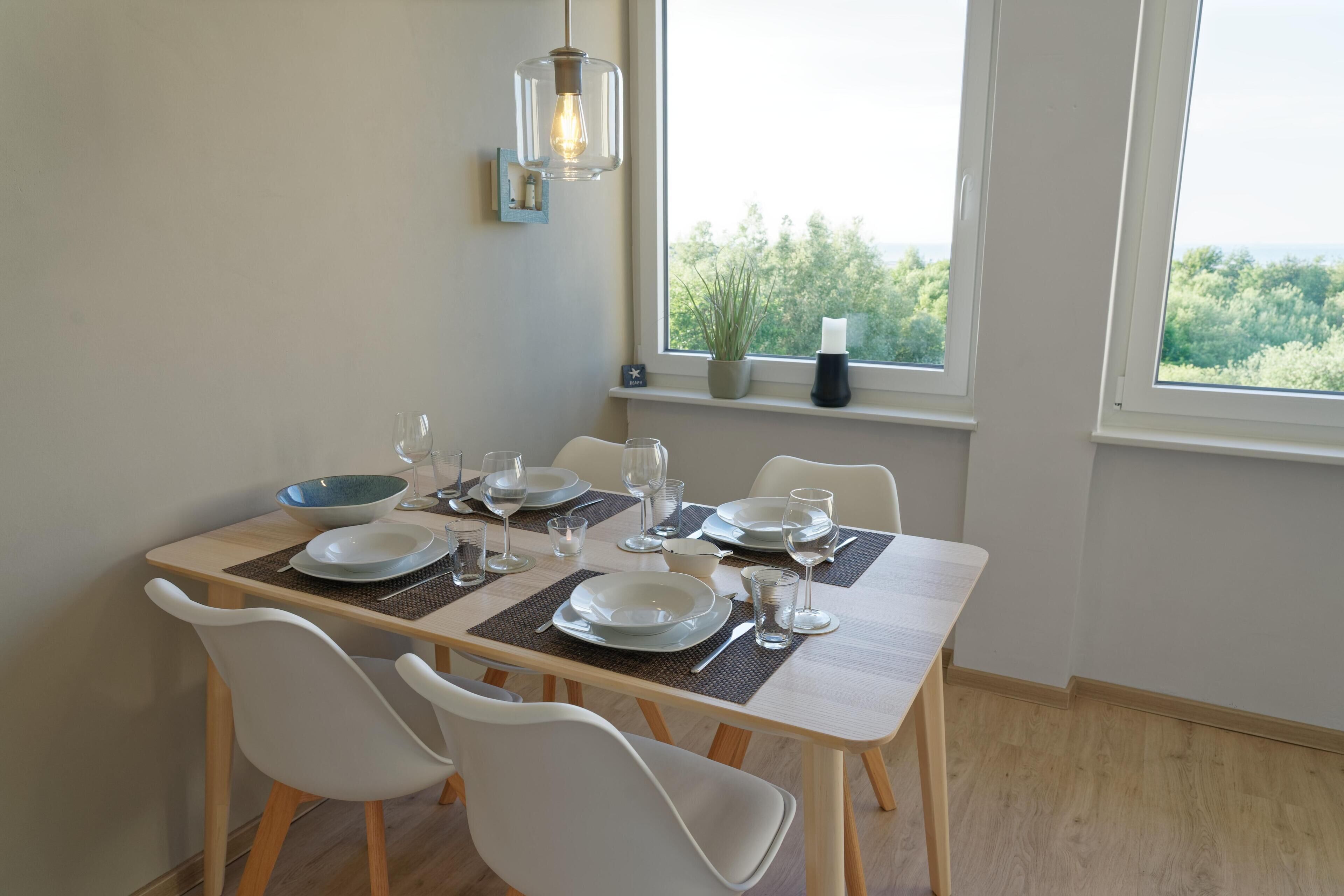 Mesa de jantar posta com pratos, copos e talheres, posicionada perto de uma janela com vista para o verde.