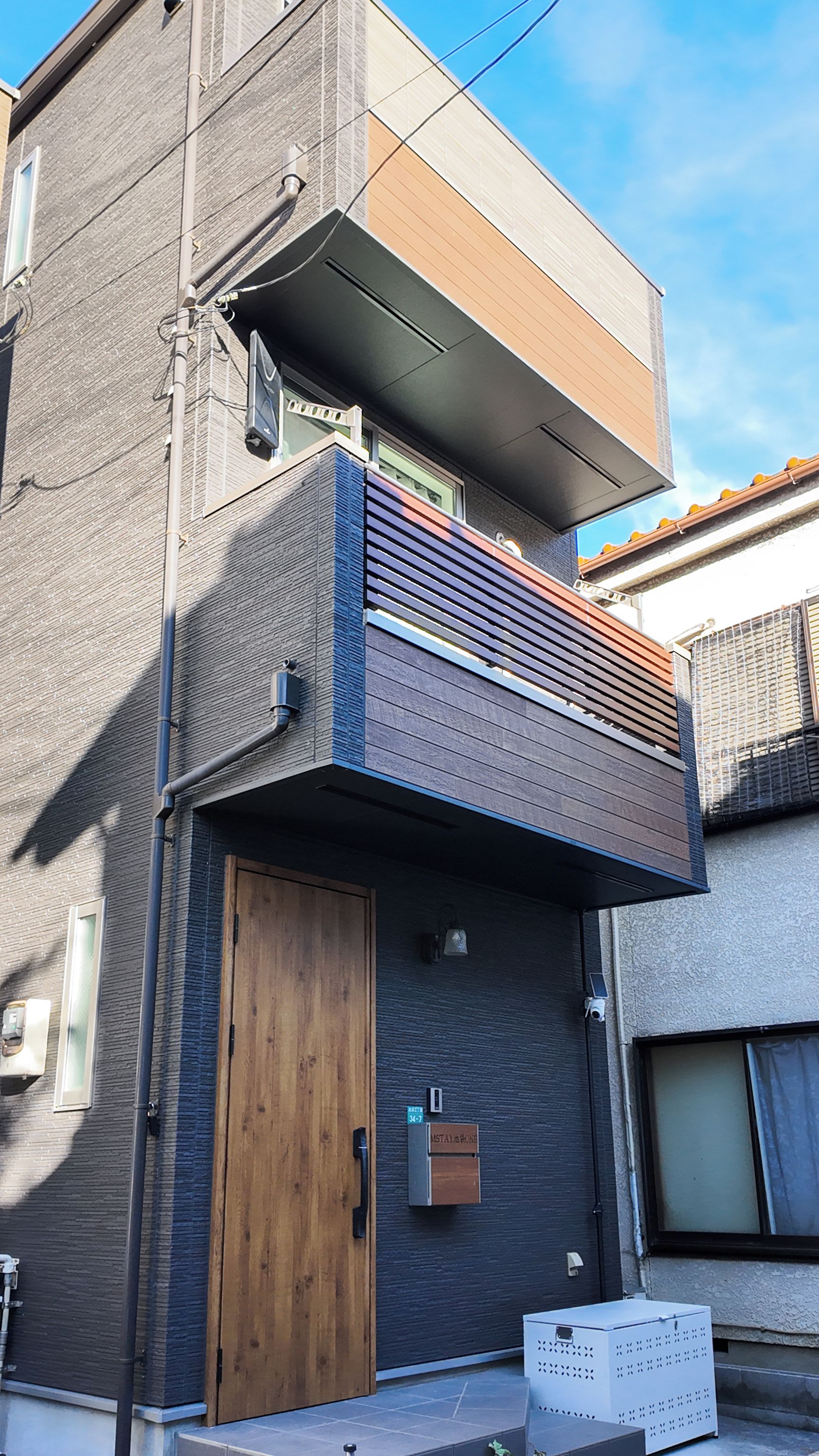A modern two-story building with a wooden door, a small balcony with a railing, and a visible mailbox.