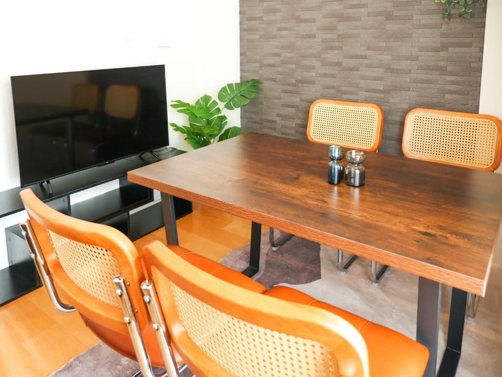 A dining area with a wooden table, two orange chairs with woven backs, and a television on the wall.