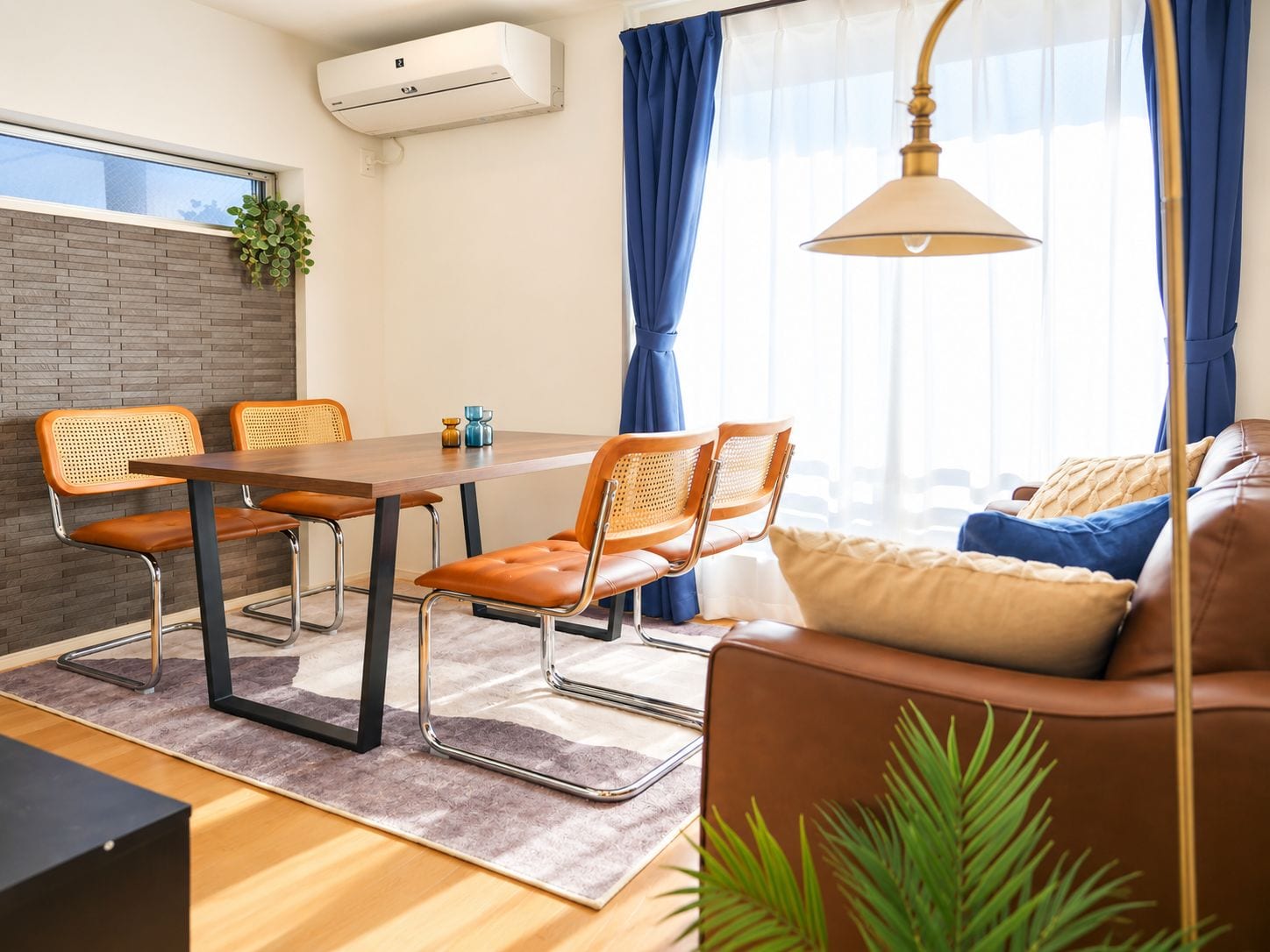 A modern living room with a dining area, featuring a brick wall, a large window with blue curtains, a sofa, and a table with chairs.