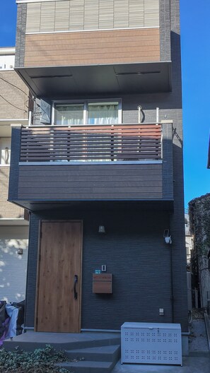 Un edificio moderno de dos pisos con puerta de madera, buzón y un pequeño espacio con mesas y sillas al aire libre.