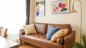 A brown leather sofa with cushions, a dining table, a lamp, and two abstract paintings on the wall.