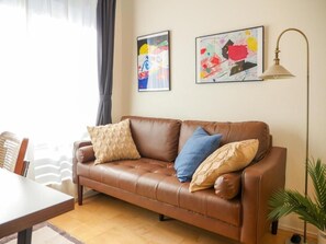 A brown leather sofa with cushions, a dining table, a lamp, and two abstract paintings on the wall. - MSTAY Ikebukuro One (Toshima-ku)