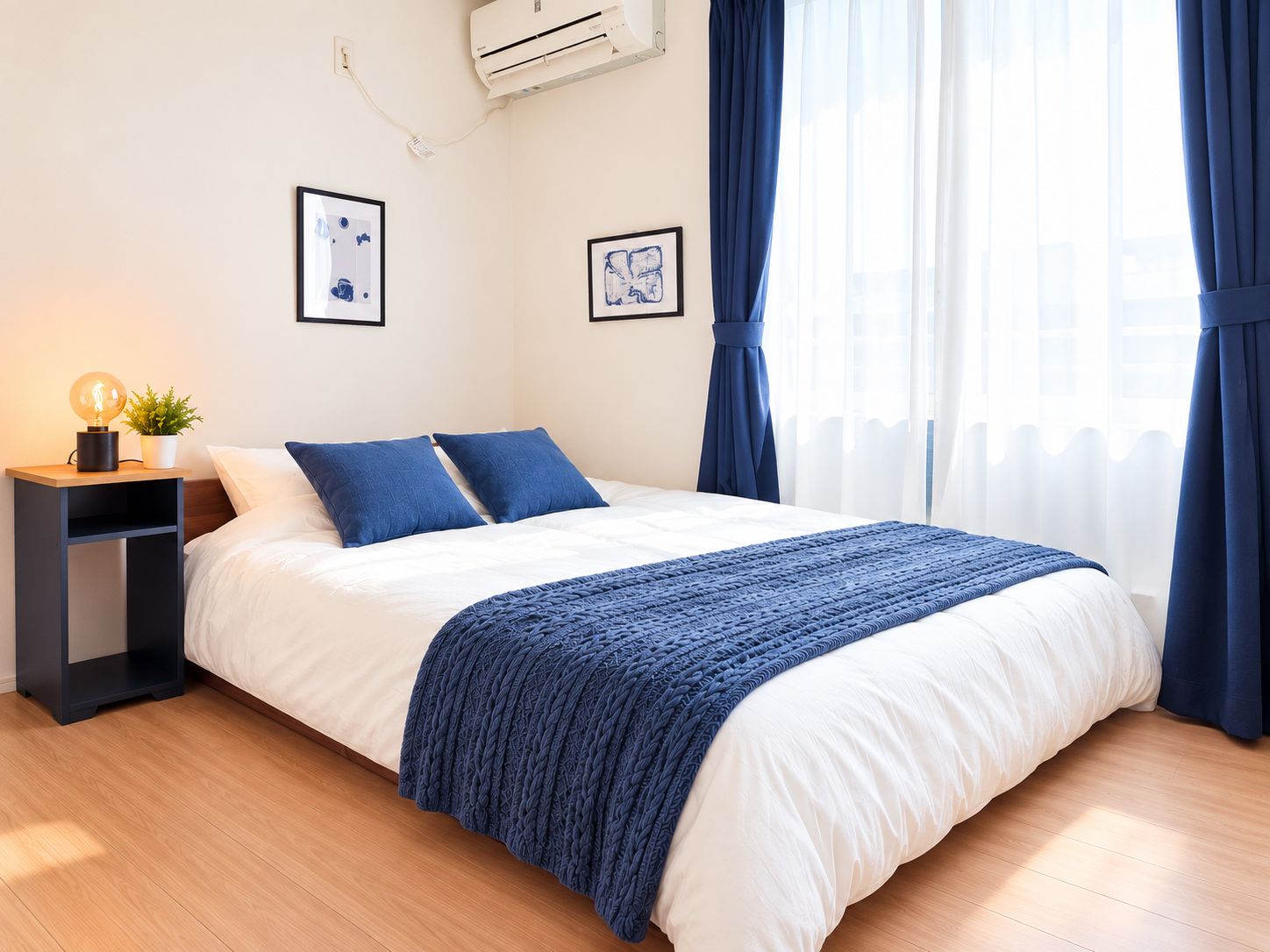 A neatly made bed with a white and blue bedspread, a bedside lamp, and a small plant on a nightstand.
