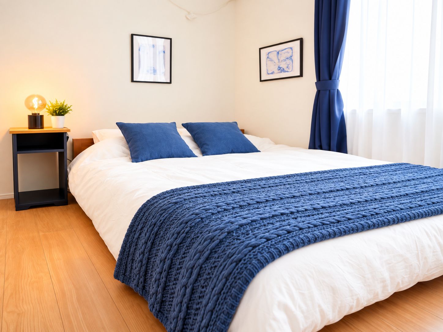 A neatly made bed with a blue blanket, a bedside table with a lamp and a plant, and two framed artworks on the wall.