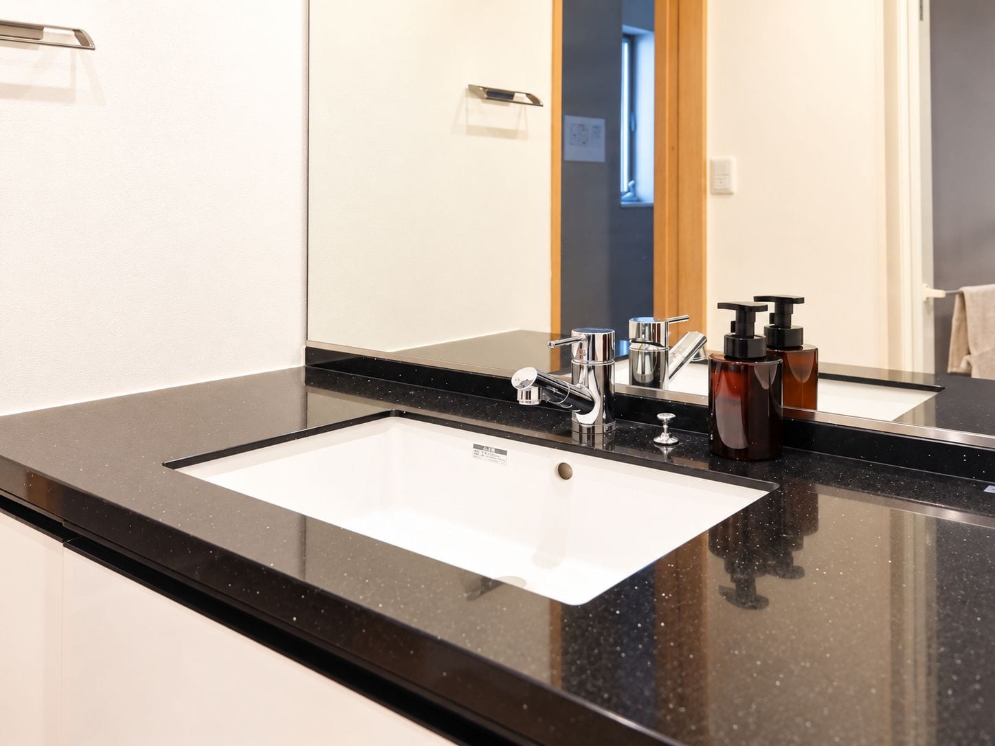 Un baño moderno con cubierta negra, un lavamanos blanco y un espejo que refleja una puerta y una ventana.