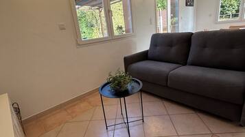 Un salon avec un canapé foncé, une petite table ronde ornée d’une plante et de grandes fenêtres donnant sur un paysage verdoyant.