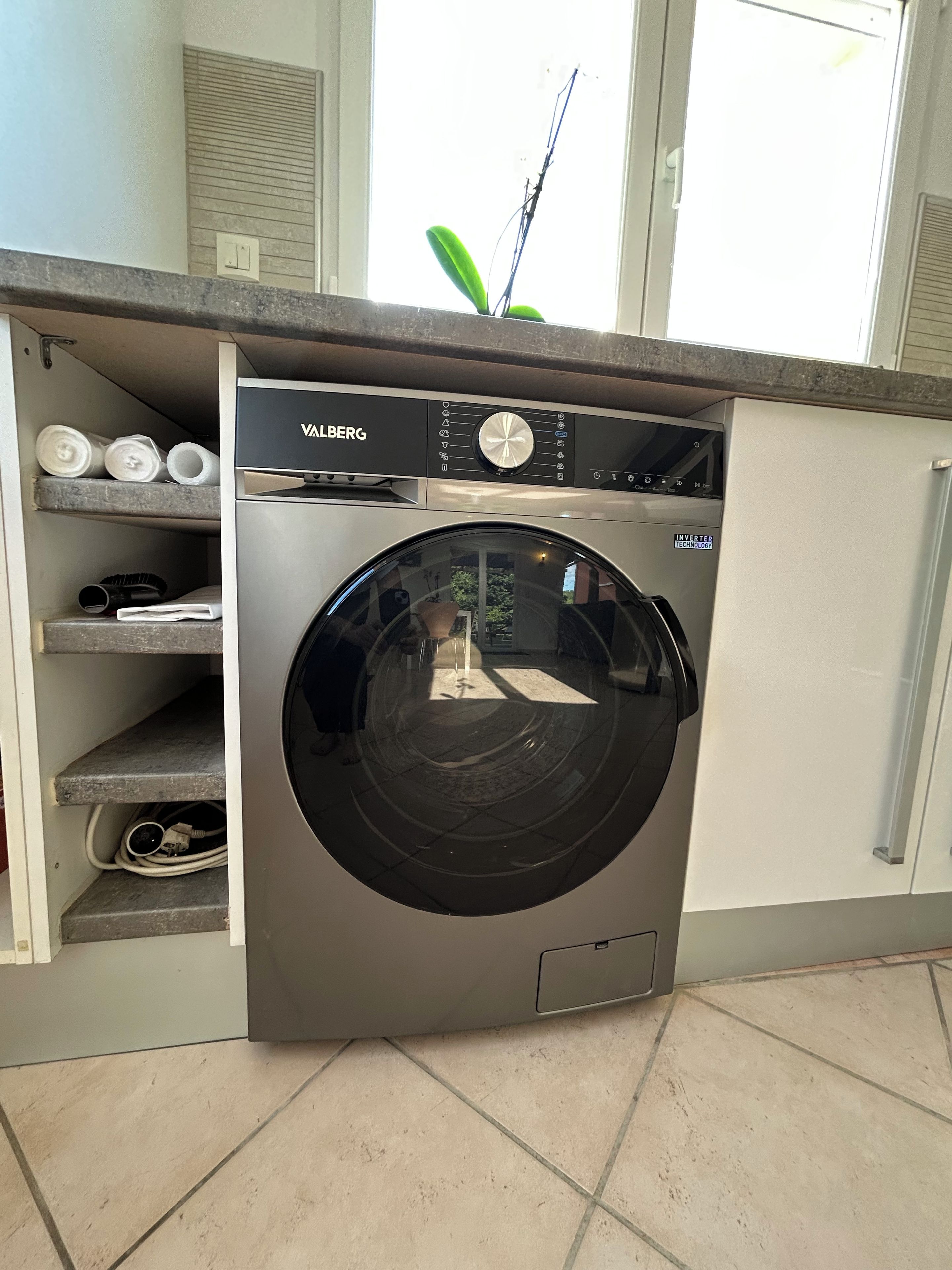 A compact, modern laundry room with a built-in washer and dryer, a small shelf with rolled-up items, and a window above.
