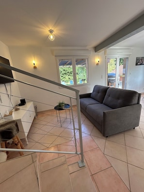 Ein modernes Wohnzimmer mit einem grauen Sofa, einer Treppe, einem Fernseher und einem Fenster mit Blick auf Grünflächen.