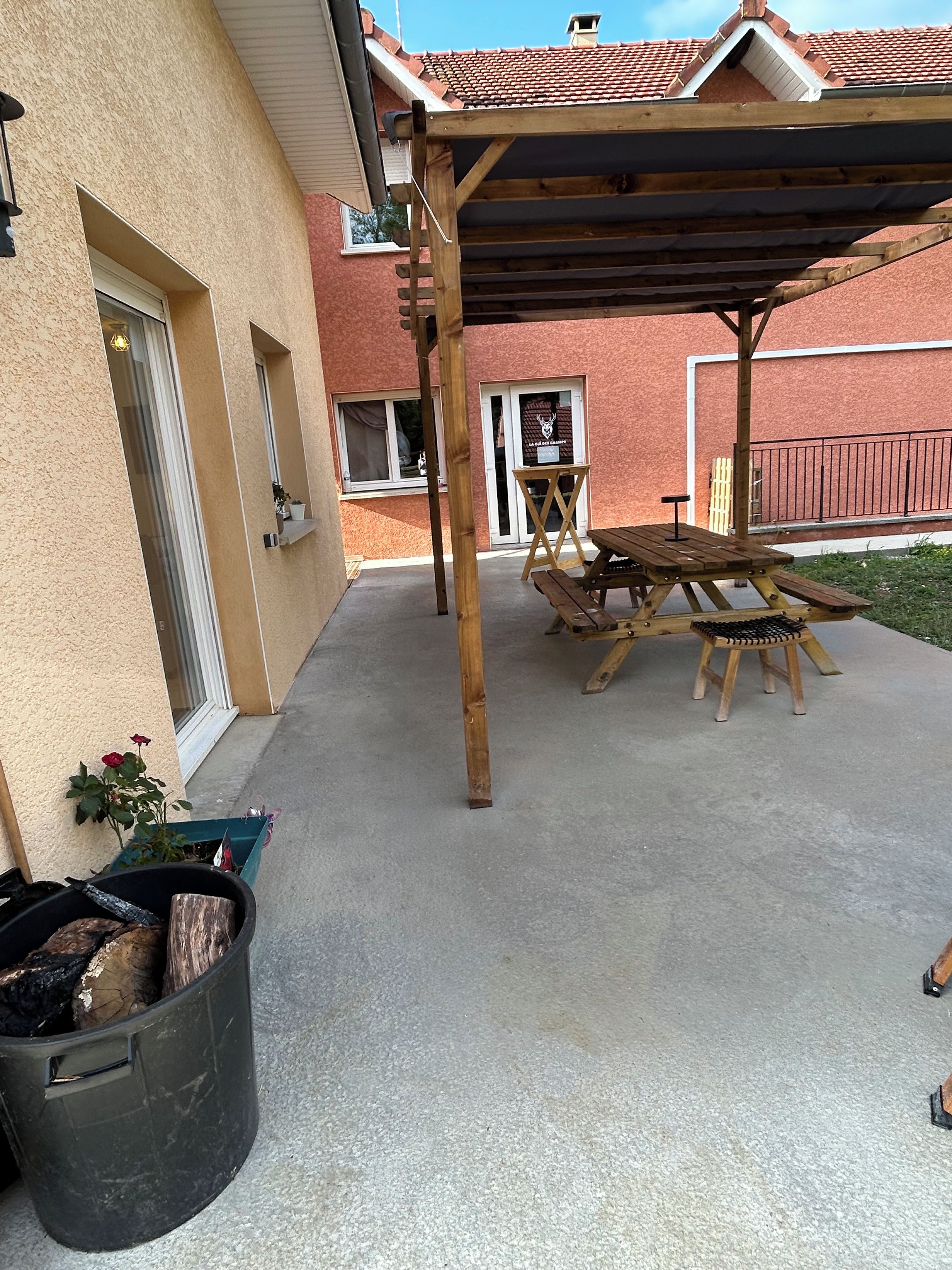 A covered patio area with a wooden table and chairs, a black trash bin, and a potted plant.