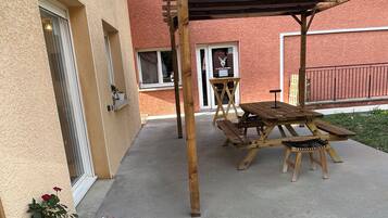 A covered patio area with a wooden table and chairs, a black trash bin, and a potted plant.