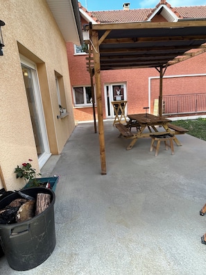 Ein überdachter Patio-Bereich mit Holztisch und -stühlen, einer schwarzen Mülltonne und einem Blumentopf.