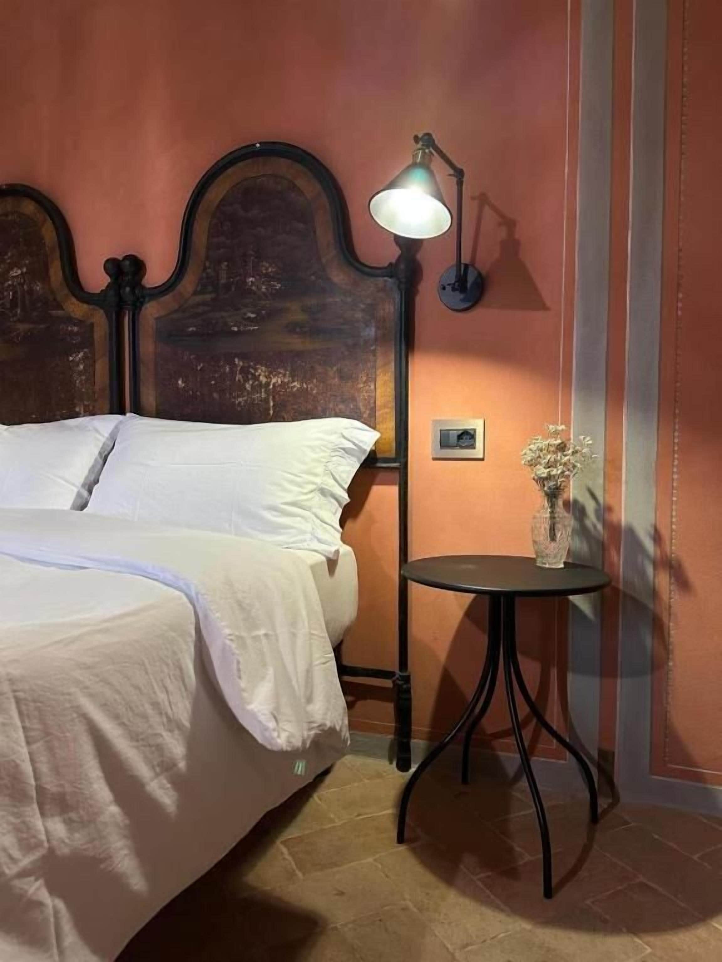 Ein Bett mit Kopfteil, ein Nachttisch mit Lampe und eine kleine Vase mit Blumen.