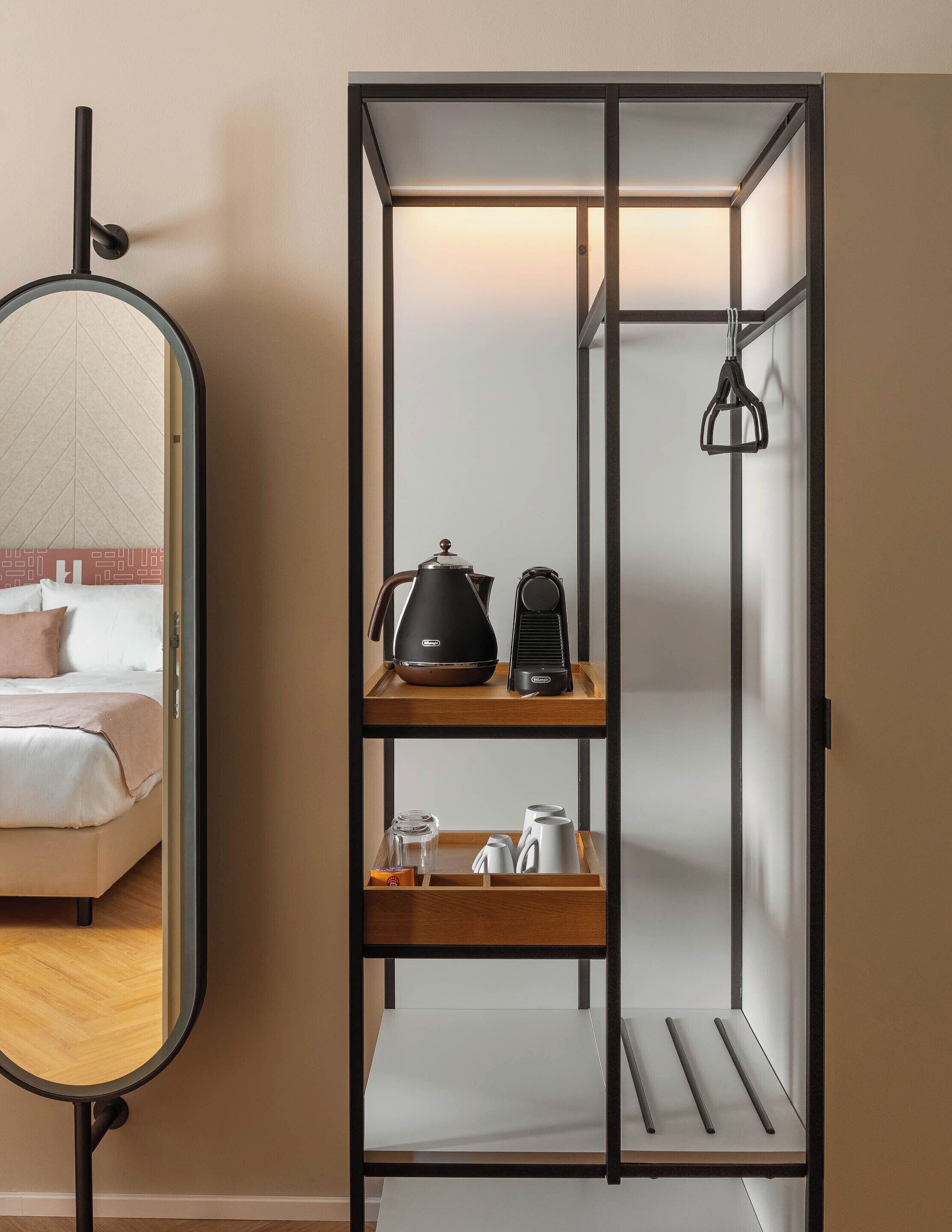 A compact room with a bed, a mirror, a coffee maker, and a shelf with cups and a kettle.