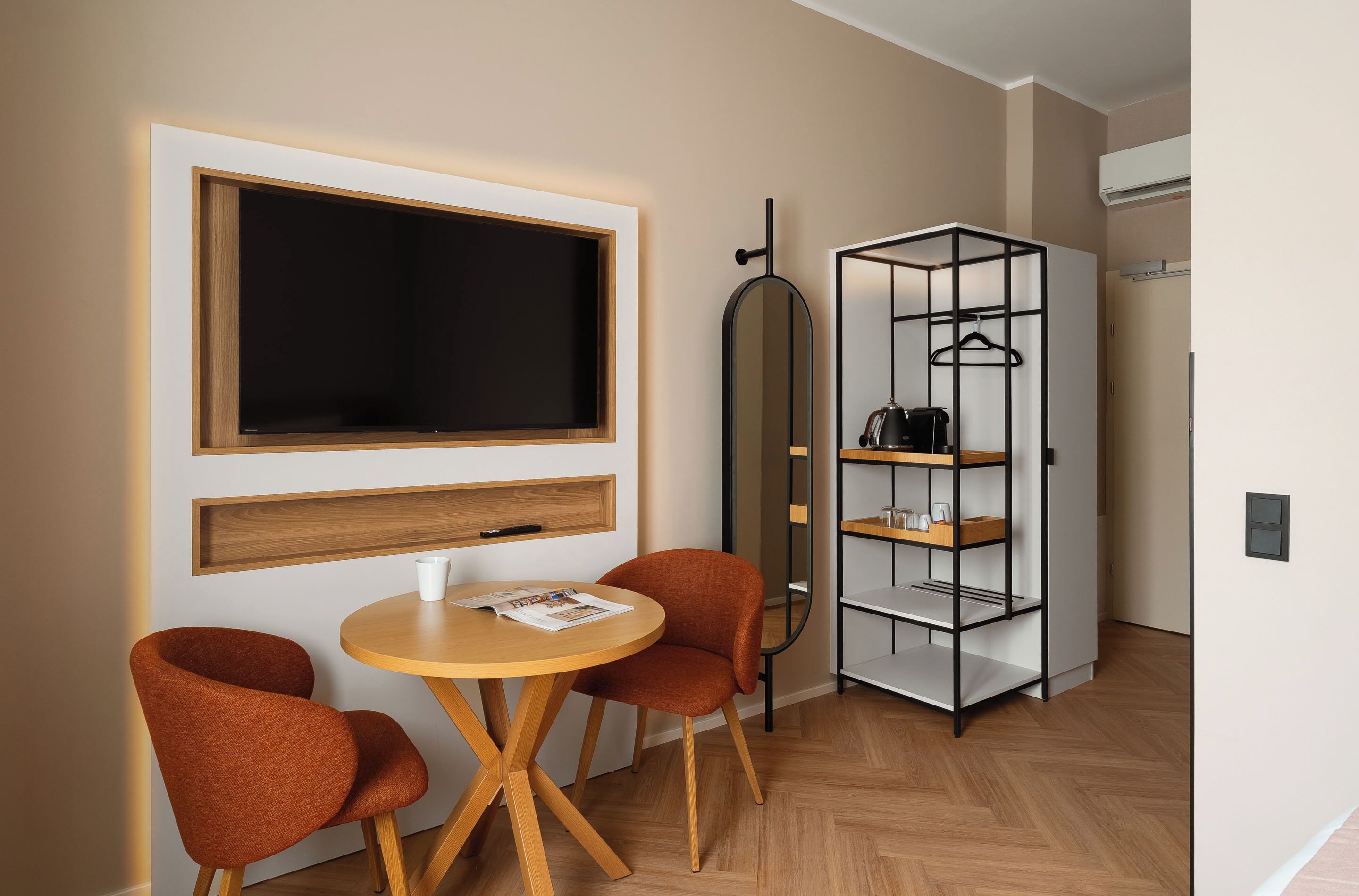 A modern room with a large flat-screen TV, two orange chairs, a round wooden table, and a black-framed mirror.