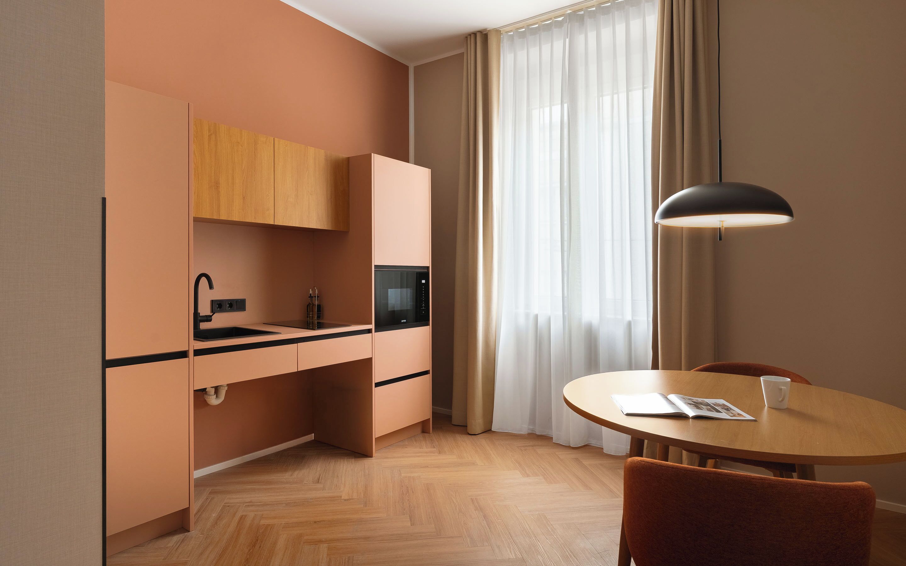 A compact kitchenette with a sink, microwave, and cabinets, a dining table with a chair, and a large window with sheer curtains.