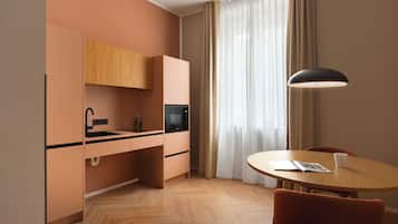 Una kitchenette funcional con lavamanos, microondas y armarios, una mesa de comedor con una silla y un ventanal con cortinas translúcidas.