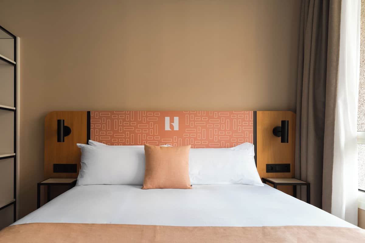 Una habitación de hotel con una cama, dos mesitas de noche, cabecero con estampado geométrico y una ventana con cortinas.