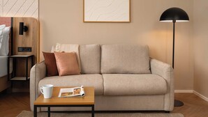 A living room with a sofa, a coffee table, a magazine, a lamp, and a wall-mounted artwork. - Limehome Linz Urfahr (Linz)